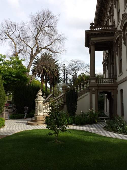 A grand mansion with ornate architecture, surrounded by lush greenery and palm trees, under a cloudy sky.
