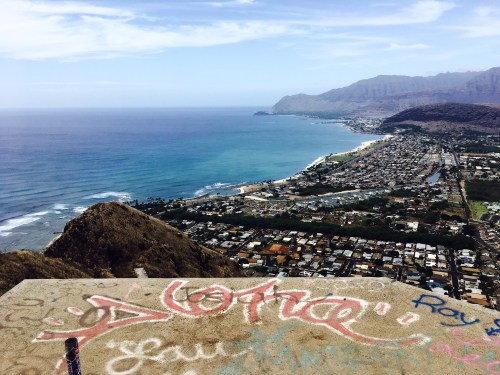 A scenic view of a coastline with mountains, ocean waves, and a city below, featuring colorful graffiti in the foreground.
