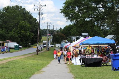 A lively street scene with people walking past colorful vendor tents at a fair on a sunny day.