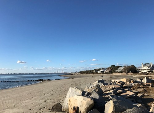 A sandy beach with large rocks in the foreground, lined by houses under a clear blue sky.