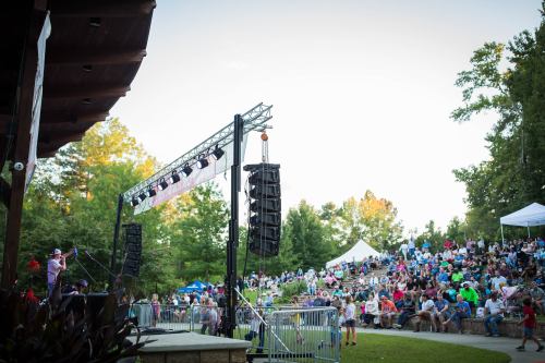 A lively outdoor concert scene with a crowd gathered on grassy steps, enjoying a performance on stage.