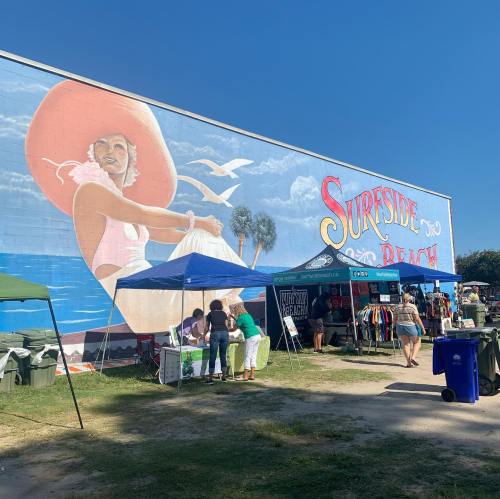 A vibrant mural of a woman in a large hat on a beach wall, with people browsing tents at a market below.
