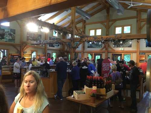 A busy winery tasting room with people mingling, wooden beams, and wine bottles on display.