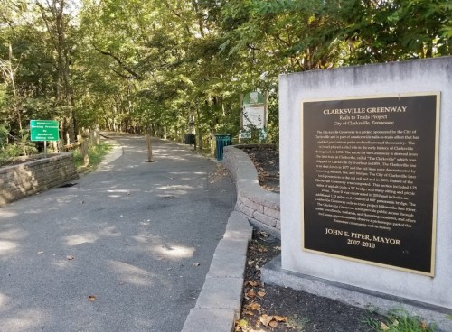 A paved path through a wooded area, with a plaque about the Clarksville Greenway project on the right.