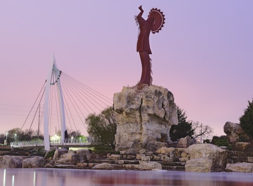 A large statue on a rock at sunset, with a modern bridge in the background and calm water in the foreground.