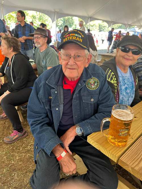An elderly man in a veteran cap sits at a table with a beer, surrounded by people at an outdoor event.