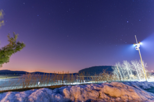 A serene night scene with a starry sky, snow-covered ground, and a distant hill illuminated by a light pole.