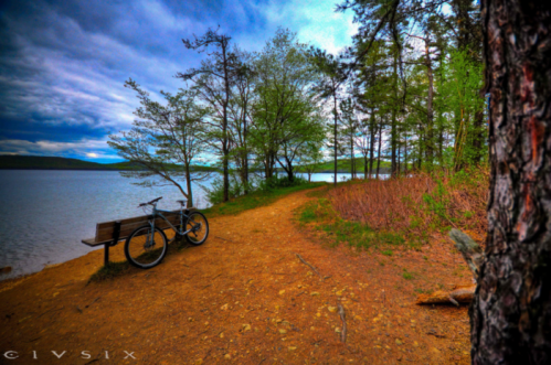 A bike rests on a bench by a serene lake, surrounded by trees and a winding dirt path under a cloudy sky.
