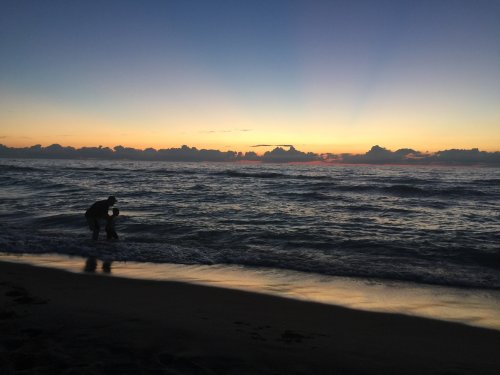 Silhouette of two people wading in the ocean at sunset, with colorful clouds and gentle waves.