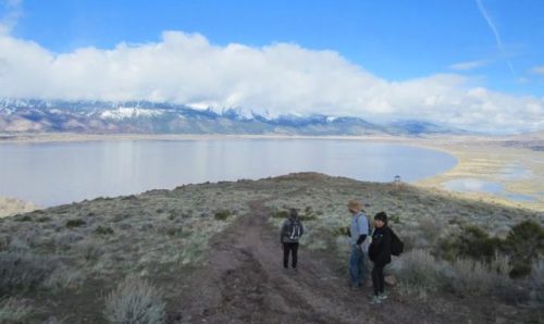 Three hikers walk along a trail overlooking a lake and snow-capped mountains under a partly cloudy sky.