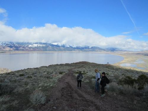 Two hikers on a trail overlooking a lake and snow-capped mountains under a partly cloudy sky.