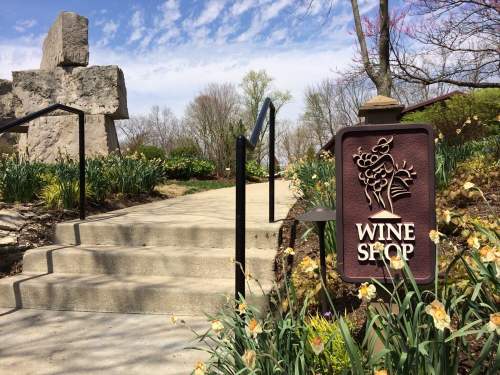 A sign for a wine shop near stone sculptures and blooming flowers, with steps leading up to the entrance.