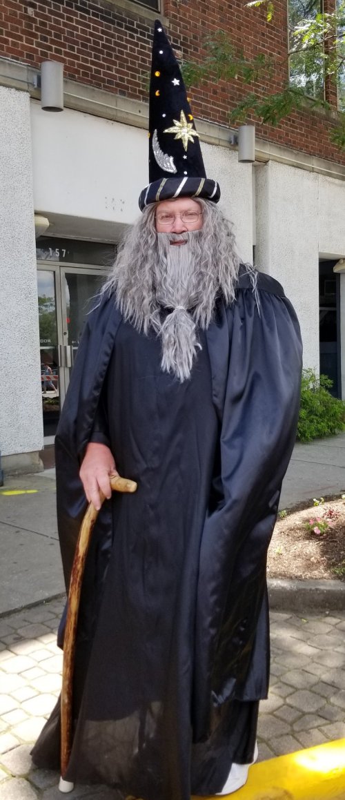 A person dressed as a wizard, wearing a black robe and a starry hat, holding a cane, standing outdoors.