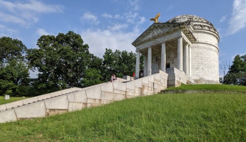 A stone monument with a dome and eagle on top, surrounded by green grass and trees, with people climbing the steps.