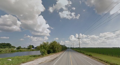 A rural road stretches into the distance, flanked by greenery and a pond under a partly cloudy sky.