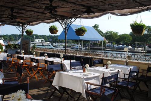 Outdoor dining area with tables set for guests, overlooking a marina with boats and greenery in the background.