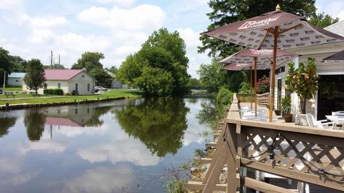 A peaceful canal scene with trees, a wooden deck, and umbrellas, reflecting clouds in the calm water.