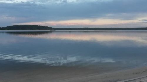A calm lake at dusk, reflecting soft clouds and a hint of color in the sky, with a sandy shore in the foreground.