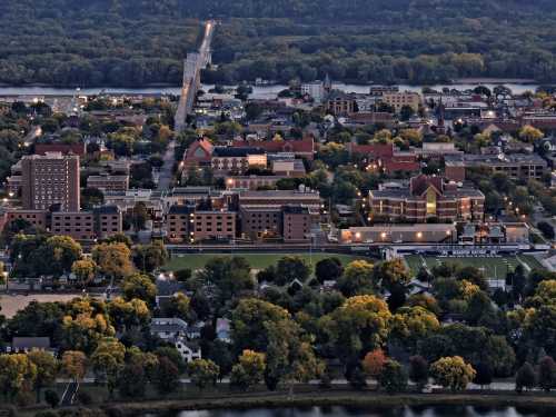 Aerial view of a town with buildings, trees, and a river, showcasing a bridge in the distance and autumn foliage.