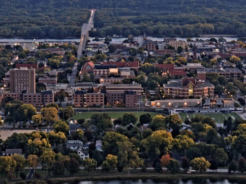 Aerial view of a town with buildings, trees, and a river, showcasing a bridge in the distance and autumn foliage.