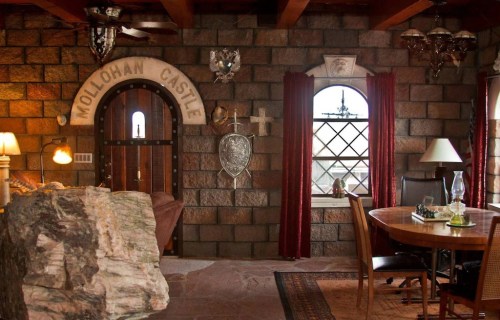 Interior of a rustic castle-themed room with stone walls, wooden beams, a round dining table, and decorative elements.