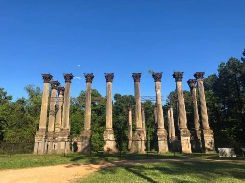 Ruins of tall stone columns against a clear blue sky, surrounded by greenery and a hint of the moon in the background.