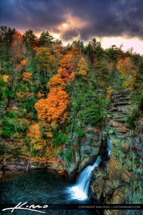 A scenic view of a waterfall surrounded by vibrant autumn foliage and rocky cliffs under a dramatic sky.