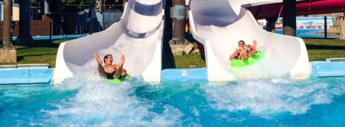 Two people sliding down water slides into a pool, creating splashes in a sunny water park setting.