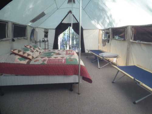 Interior of a spacious tent featuring a bed with a quilt and a folding cot, surrounded by windows and natural light.