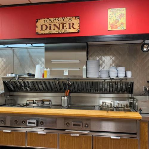 Interior of Nancy's Diner featuring a stainless steel kitchen with cooking equipment and stacked plates against a red wall.