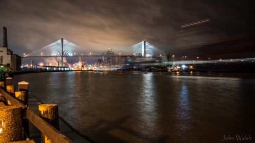 A nighttime view of a bridge over a river, with city lights reflecting on the water and clouds in the sky.