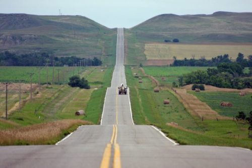 A long, straight road stretches through green fields and rolling hills, with a tractor driving in the distance.