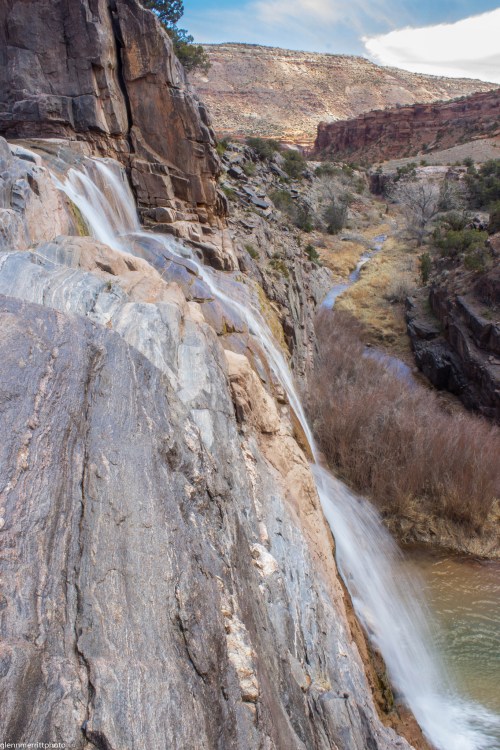 A cascading waterfall flows over rocky terrain into a serene river, surrounded by rugged cliffs and sparse vegetation.