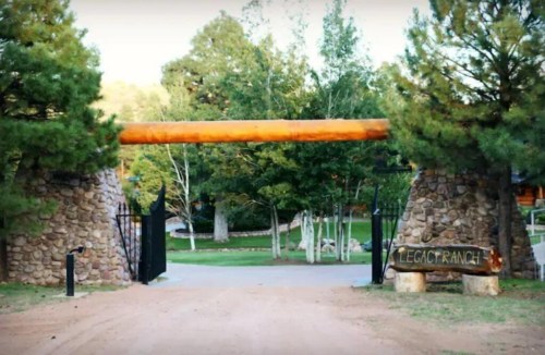 A rustic stone entrance gate with a wooden beam, leading into a lush, green ranch area.