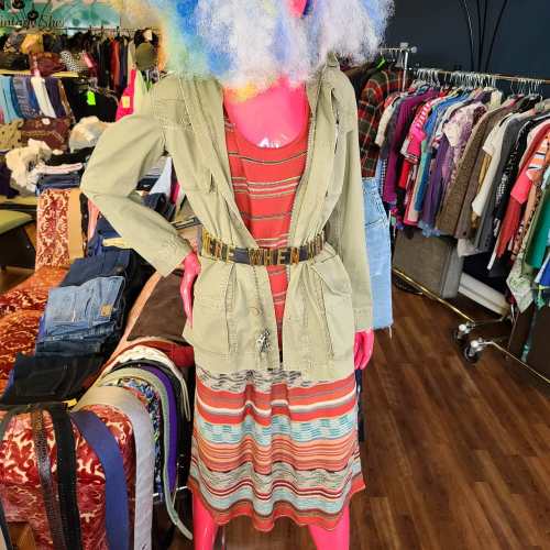 A mannequin displays a layered outfit with a green jacket over a striped dress, surrounded by colorful clothing racks.