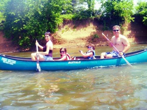 A family of four paddles a canoe on a river, surrounded by lush greenery and a sandy bank.