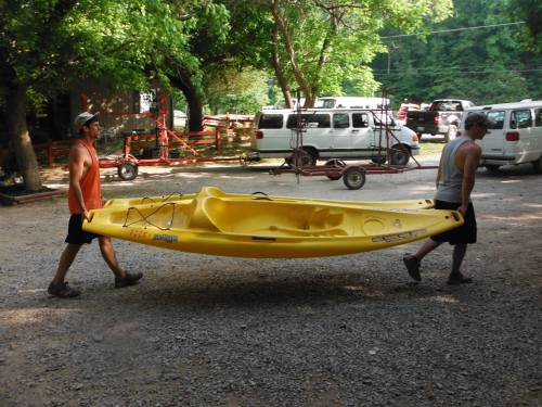 Two people carry a yellow kayak through a gravel area, surrounded by trees and parked vehicles.