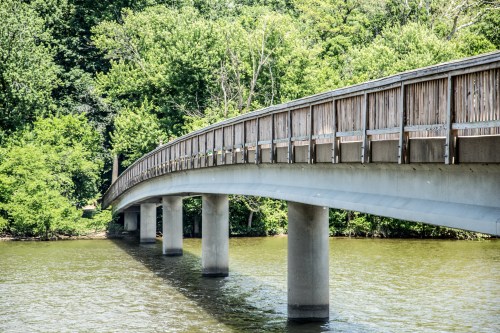 A wooden bridge arches over a calm river, surrounded by lush green trees.
