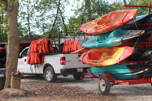 A truck and trailer loaded with colorful kayaks and life jackets, parked in a wooded area.