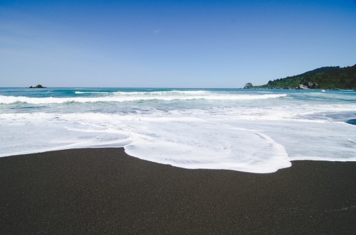 A serene beach scene with gentle waves lapping at a dark sand shore under a clear blue sky.
