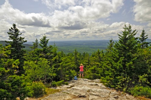 A person stands on a rocky outcrop, overlooking a lush green valley and distant mountains under a cloudy sky.