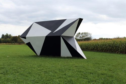 A large, abstract black and white geometric sculpture in a grassy field with trees in the background.