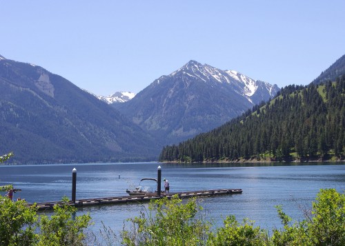 A serene lake surrounded by mountains, with a dock and a person near a boat under a clear blue sky.