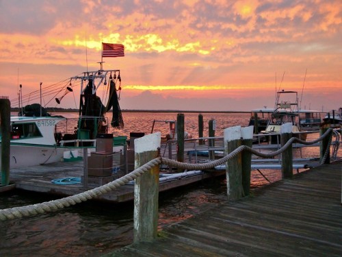 A serene sunset over a marina, with fishing boats docked and an American flag waving in the background.