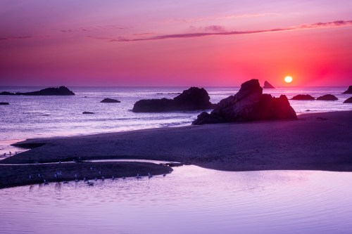 A serene beach at sunset, with vibrant purple and orange skies, rocky formations, and calm water reflecting the colors.