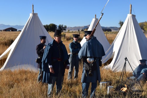 Civil War reenactors in period uniforms gather near white tents in a grassy field, with mountains in the background.