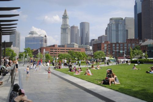 A city park with families enjoying the day, children playing in fountains, and skyscrapers in the background.