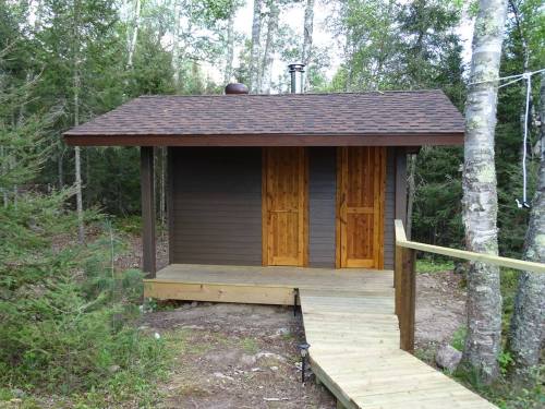 A small wooden cabin with a sloped roof, two wooden doors, and a pathway surrounded by trees.