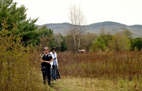 Two women walking on a path through a grassy area with trees and mountains in the background.