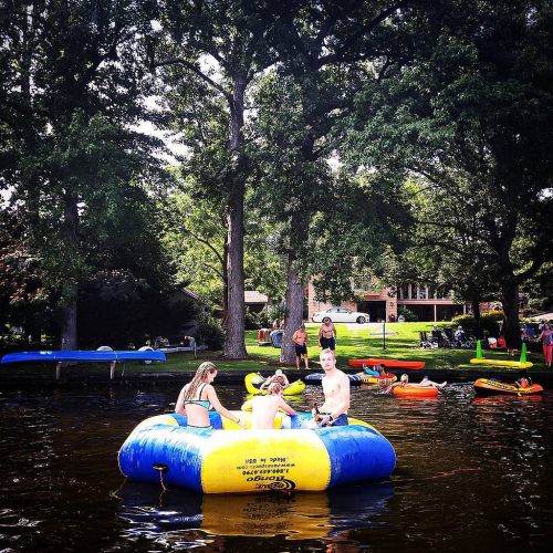 Three people sit on a blue inflatable raft in a lake, surrounded by trees and other colorful floats.